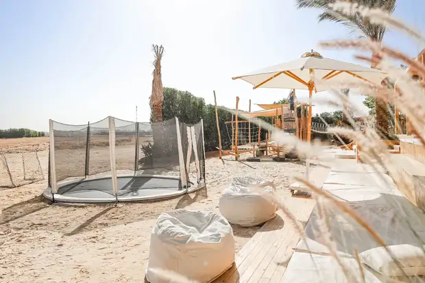 Venkovní dětské hřiště v SALT El Gouna — trampolína zapuštěná v písku, sedací vaky a slunečníky pod palmou